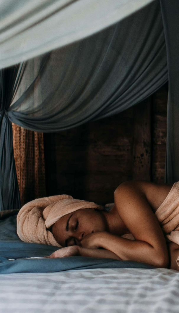 Femme se reposant après un moment de détente, allongée sur un lit entouré de rideaux drapés, illustrant un sommeil apaisant et une ambiance bien-être naturelle.