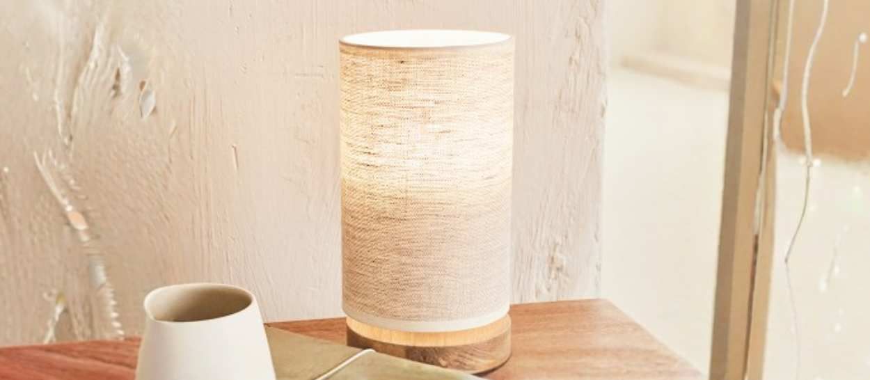 Lampe à poser en lin Kipli diffusant une lumière naturelle sur une table de chevet en bois dans une chambre minimaliste.